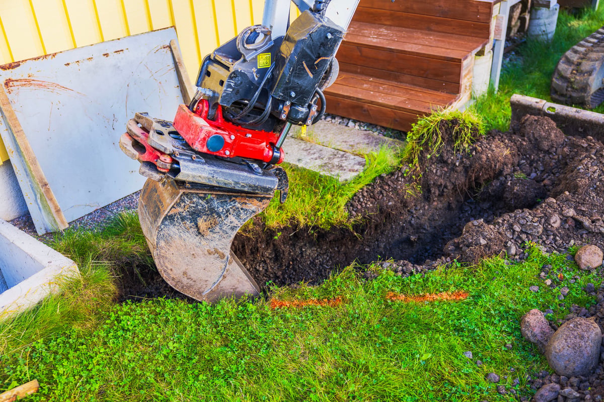 Excavator bucket digging a soil trench beside a house in an open yard