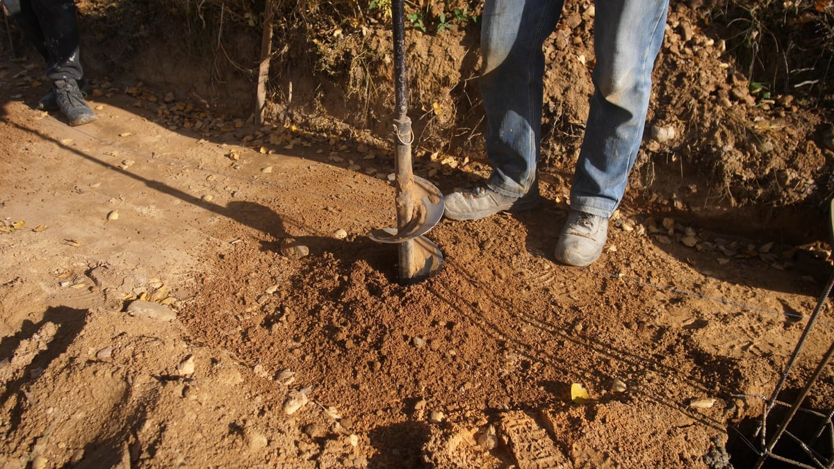 Worker using an auger to install fence posts in open, dusty terrain