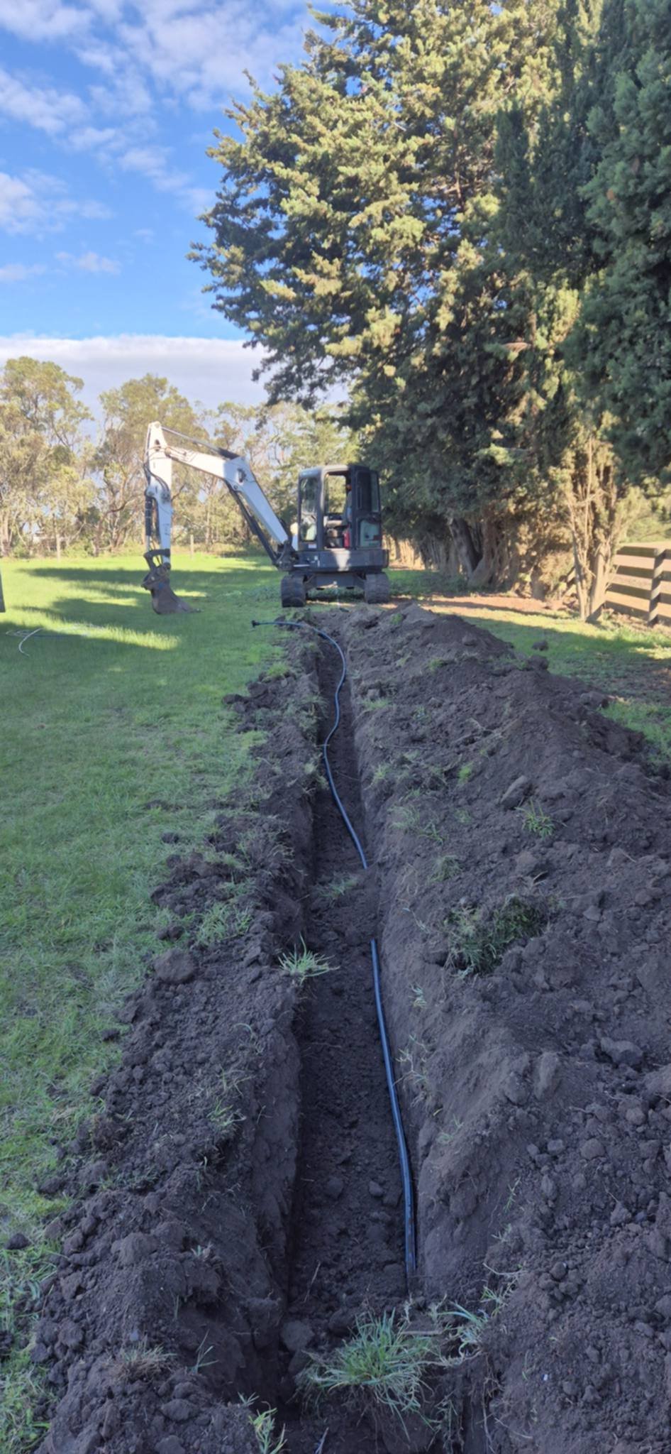 Small excavator digging a trench in a grassy field with blue irrigation pipe laid in the ground