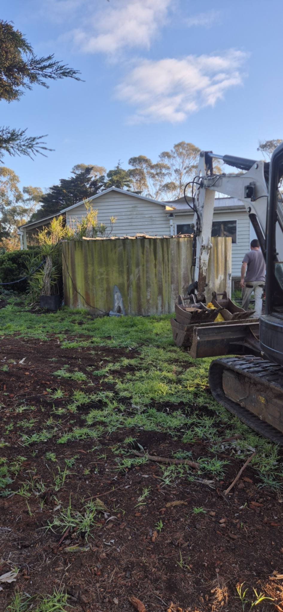 Excavator parked in the yard of a residential property with a weathered house and mossy fence