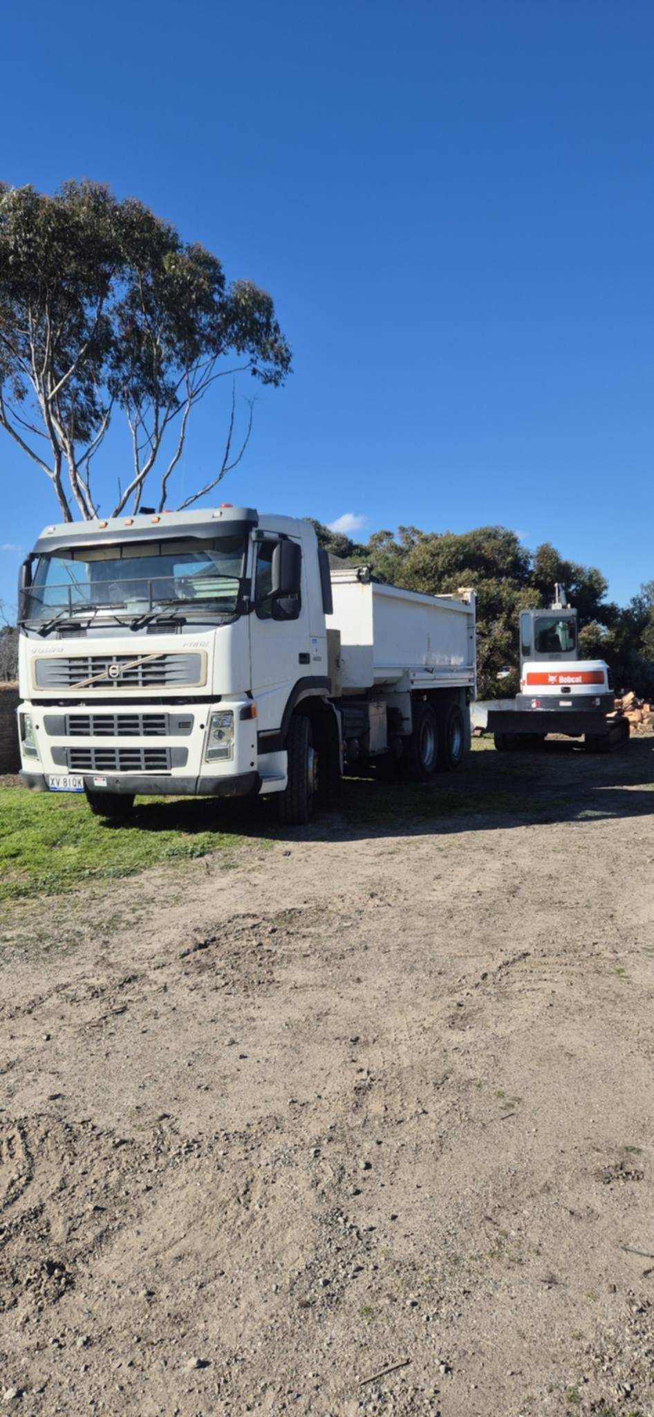 White Volvo dump truck and Bobcat excavator parked on site under a clear blue sky