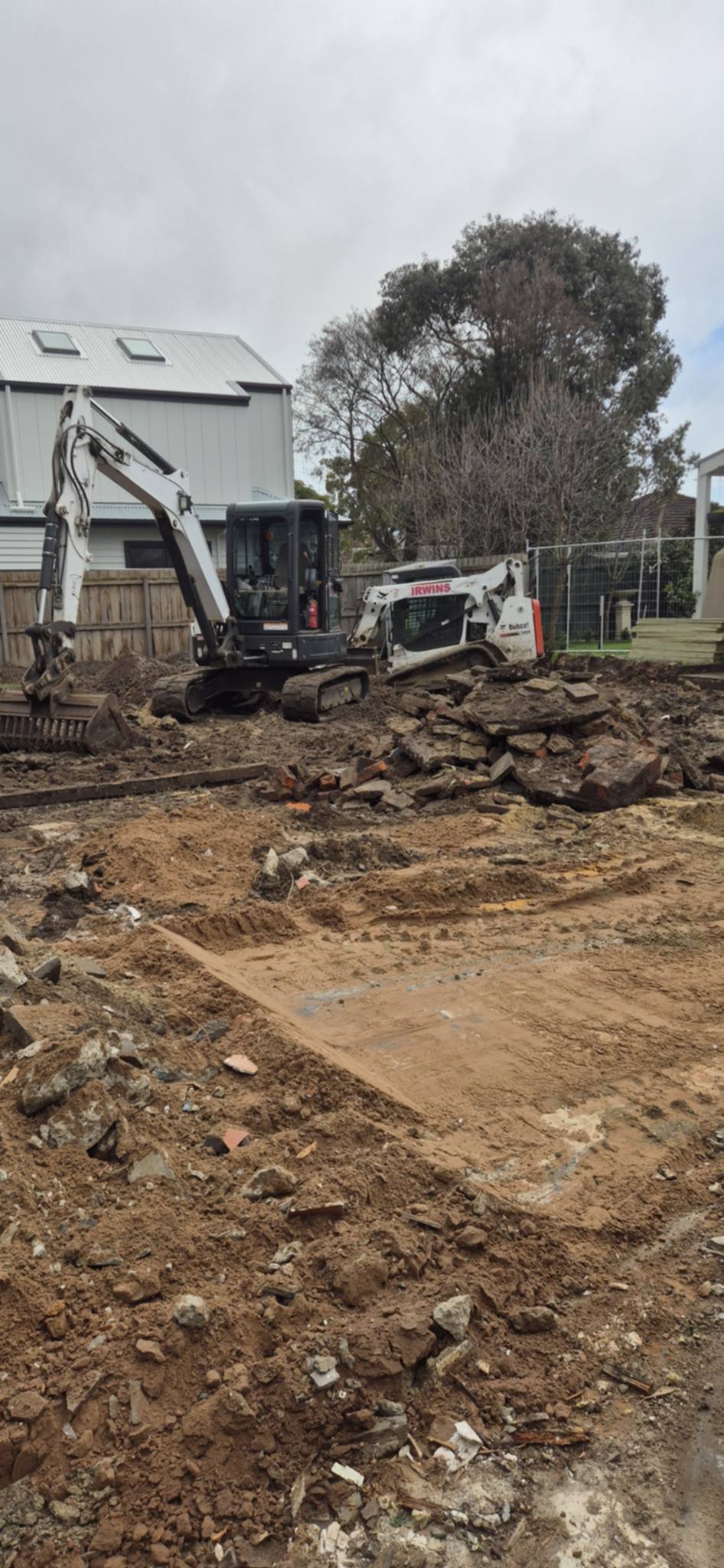 Mini excavator and Bobcat loader working a residential construction site with dirt, rocks and a metal shed in the background