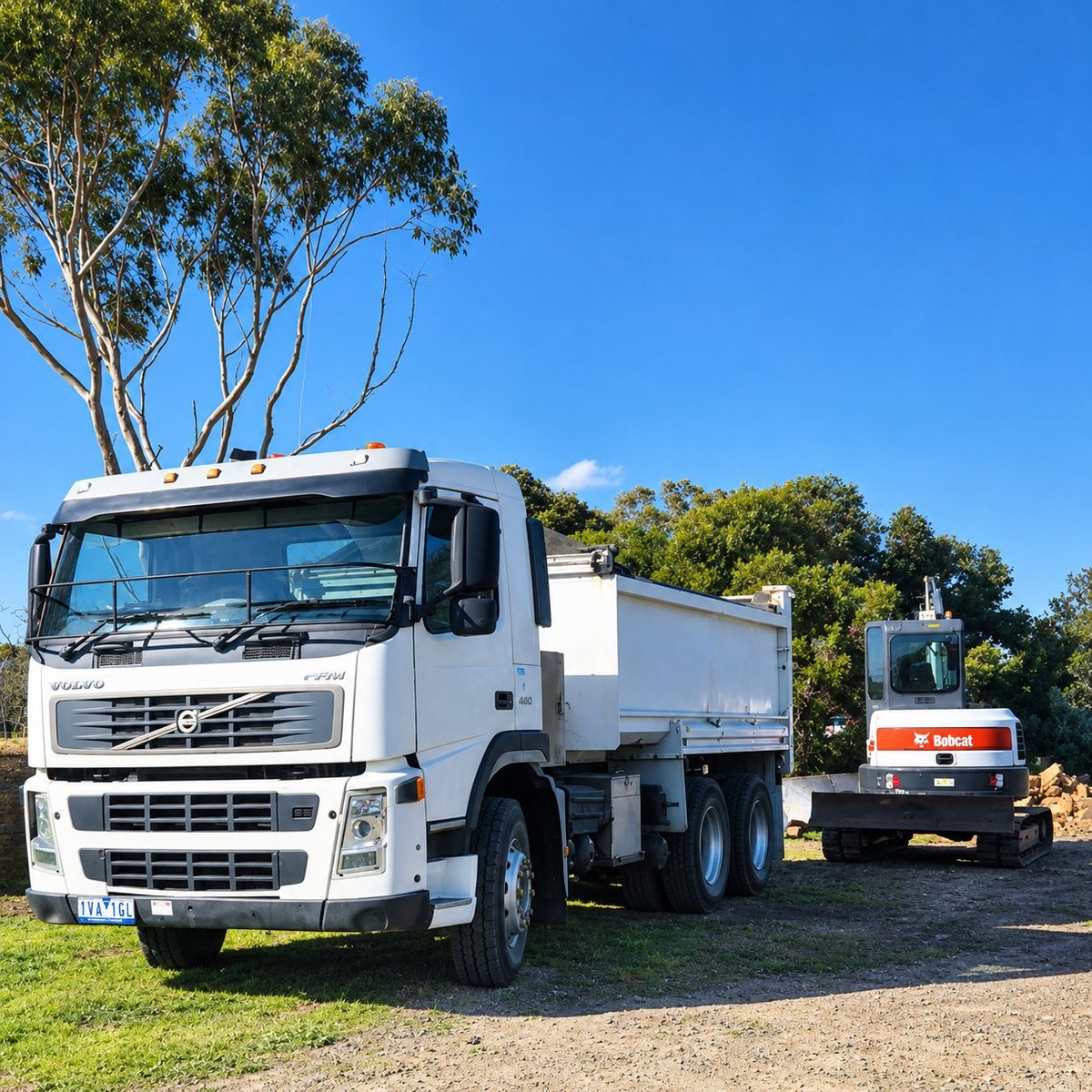 White Volvo tipper truck and Bobcat excavator parked on a Geelong job site under a clear blue sky