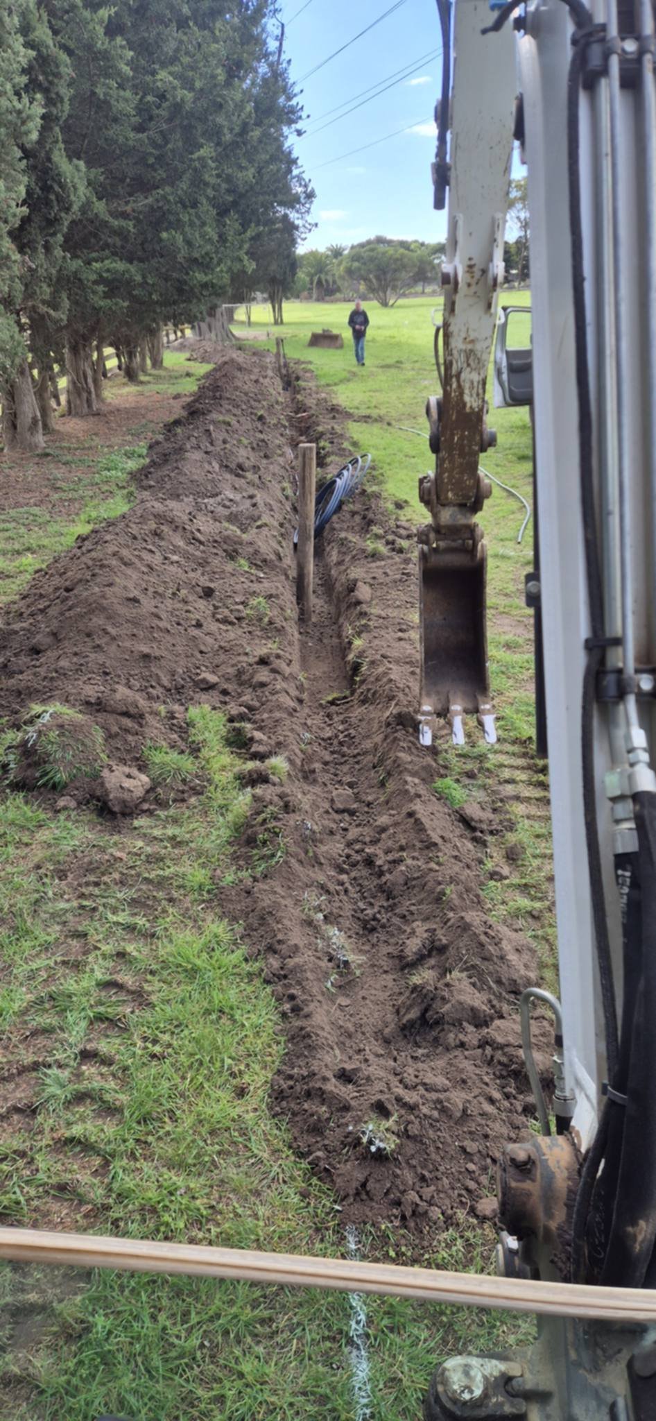Excavator bucket digging a deep trench through a grassy field with trees