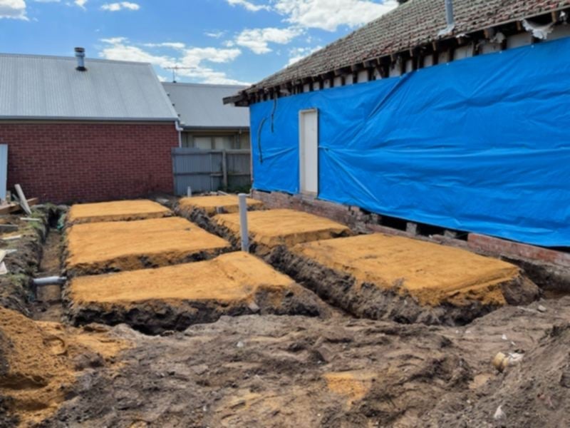 Excavated foundation sections with metal support post beside a brick building and blue-tarped structure