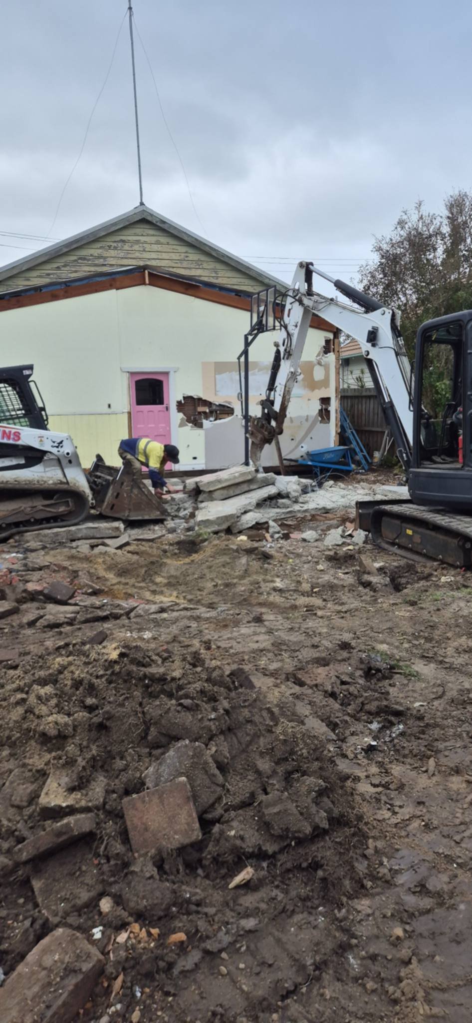 Yellow house undergoing renovation with an excavator and skid loader removing debris on muddy ground